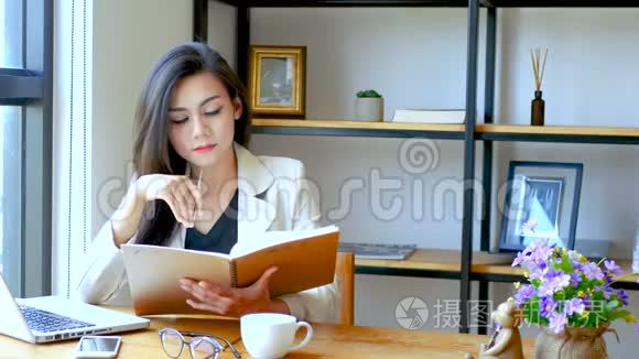 4K镜头，美丽的亚洲商务女性坐在电脑笔记本电脑前，阅读并翻翻笔记簿页面