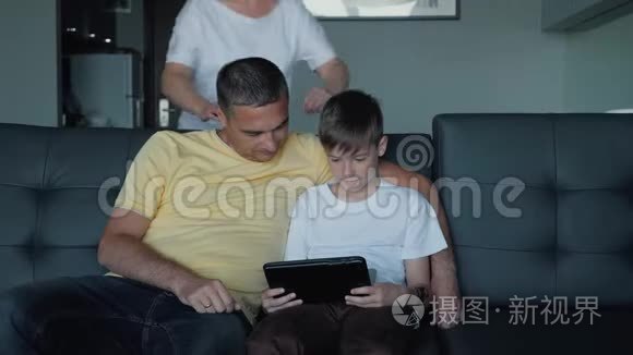 父亲、母亲和儿子坐在沙发上看电影，在平板电脑上玩游戏。 一个幸福的家庭