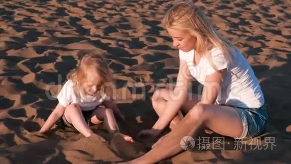 美丽的金发妈妈和女儿坐在海滩上玩沙子。