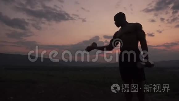 非洲裔美国人肌肉健美的身体建设者的剪影举着哑铃在粉红色日落的天空背景。 室外