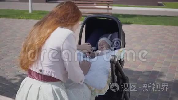 美丽的红发女人和她的孩子在公园里躺在婴儿车里玩。 这位女士和她一起享受阳光明媚的一天