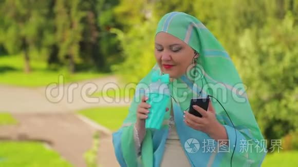 公园里一位戴着浅色围巾的年轻穆斯林妇女正在喝咖啡，用耳机在信使里说话。