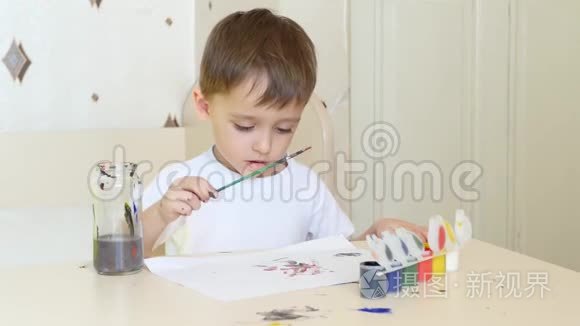一个小男孩坐在桌子旁，用画笔在一张纸上画画。 水彩颜料。