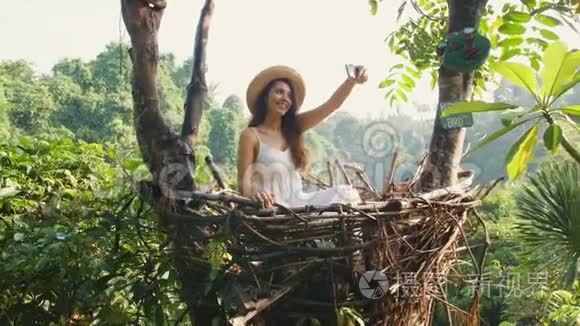 年轻的快乐混血旅游女孩穿着白色连衣裙，用手机拍摄自拍照片，坐在装饰草窝里。