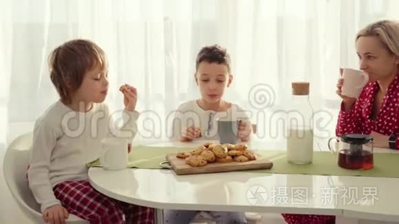 两个男孩在现代厨房吃早餐。 男孩吃带牛奶的饼干。 厨房里的白色桌子。 母亲和两个儿子