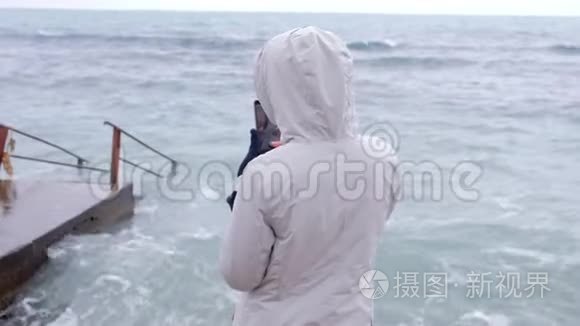 穿白色夹克的女人拿着手机风浪站在堤岸上。 后景。