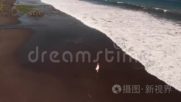 4巴厘岛黑色火山海滩上，身穿白色礼服的年轻女子行走的空中飞行镜头。