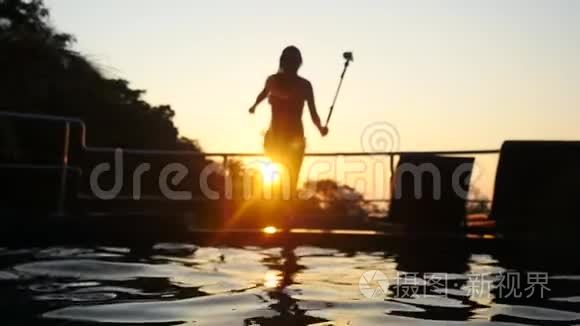 美丽的年轻女子在美丽的夕阳下在游泳池里跳着剪影，镜头闪耀