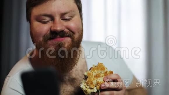 一个长着大胡子的胖子在他的智能手机里打东西，在沙发上吃汉堡