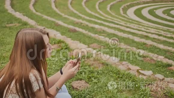 一个年轻漂亮的女孩手里拿着一部手机，坐在一个绿色的大体育场里的草地上。 娱乐。