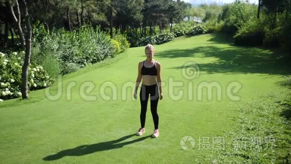 年轻的金发女人带着辫子在绿色的夏季公园的草地上做跳跃千斤顶健身锻炼。 慢慢