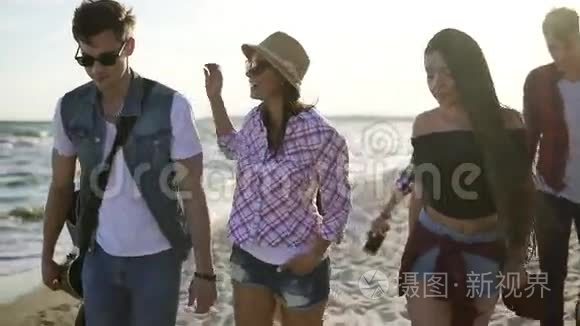 一群年轻的嬉皮士朋友一起走在海滩上，在水边`拿着吉他。 慢镜头