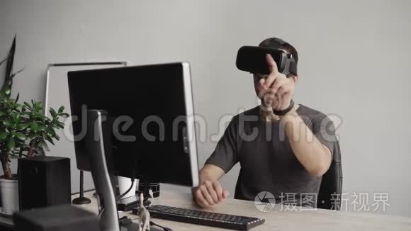 年轻人戴着虚拟现实护目镜，坐在办公室里对着电脑，试图触摸物体