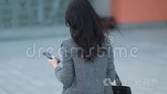 年轻的商务小姐穿着一件紧身夹克，头发深棕色，手里拿着智能手机，走在城市的大街上
