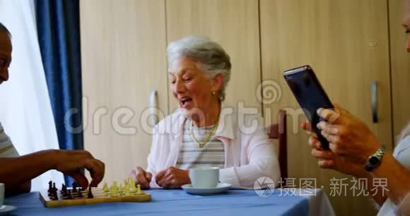 一位年长的女士使用数码平板电脑，而她的朋友正在下棋