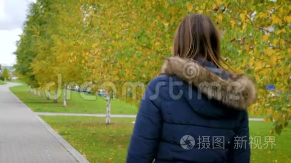 一个年轻的女人沿着城市公园的秋巷散步。 女孩走在秋日的桦树上