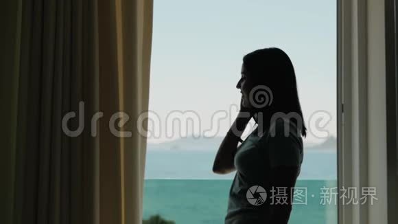 年轻女子用智能手机打电话的剪影。 高加索模特站在窗口，拿着手机