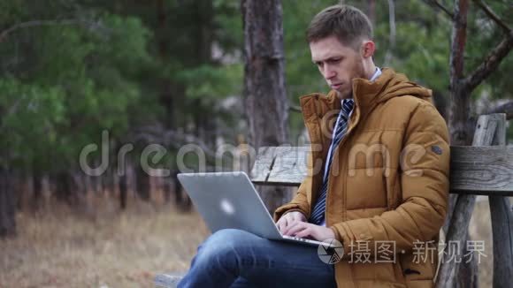 那个穿着领带和夹克的人正坐在一个秋天公园的长凳上，拿着一台笔记本电脑。 在键盘上输入文字，冲浪