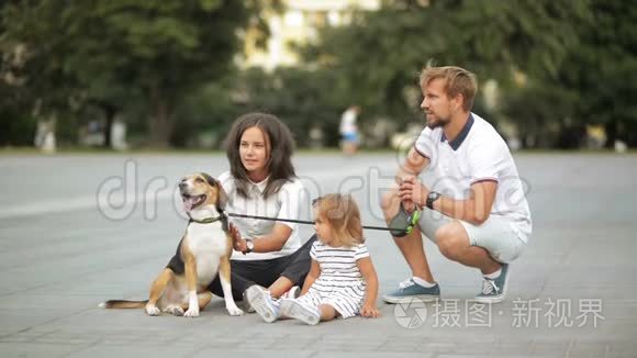 漂亮的年轻母亲留着胡子的父亲可爱的小女儿和小猎犬正坐在城市广场上