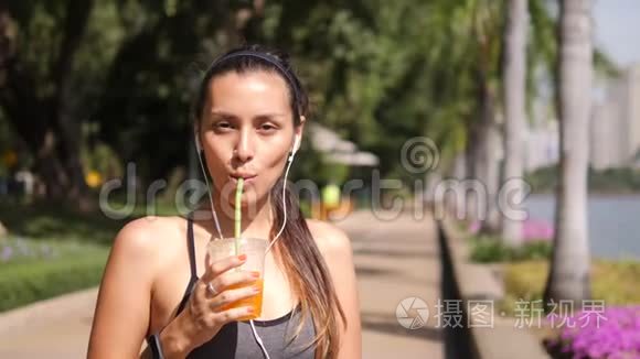 迷人的混合种族瑜伽女孩喝新鲜橙汁后，在城市公园。 4K，慢动作。 泰国。