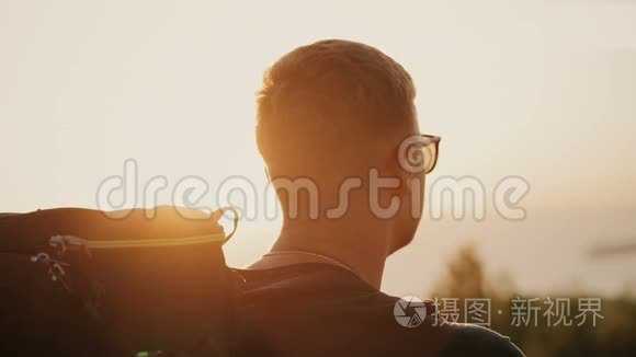 慢动作的英俊年轻时尚男子戴着太阳镜和背包看着夕阳在田野。 在一个
