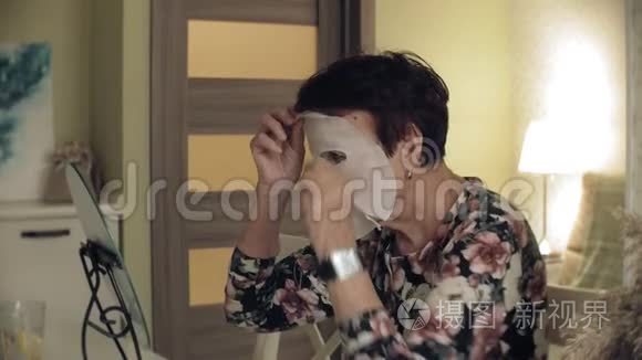 一位老妇人照镜子，从脸上摘下一个化妆组织面具。 面部护理。 整形手术和胶原蛋白