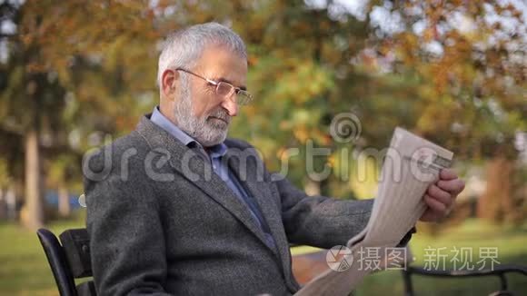 大胡子老人戴眼镜在秋天公园看报纸。 英俊的白发男子早早地坐在长凳上