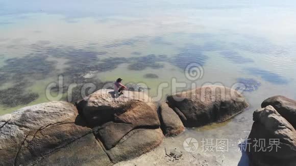 戴帽子的年轻人在海边享受自由，独自坐在岩石上。 从无人机飞到大海的视野。 1920x1080
