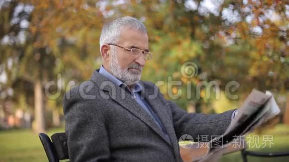 大胡子老人戴眼镜在秋天公园看报纸。 英俊的白发男子早早地坐在长凳上
