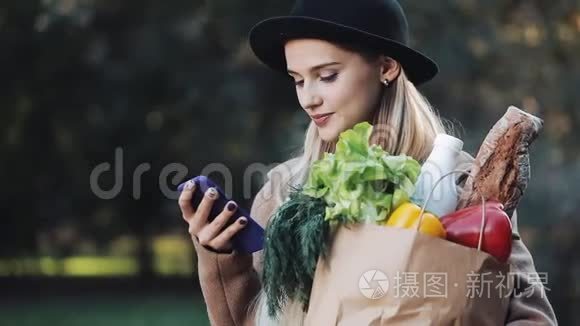 年轻漂亮的女人穿着时髦的外套，站在秋天的公园里，拿着一包产品，用智能手机