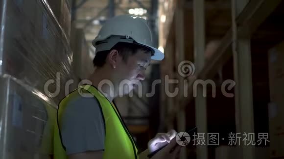 年轻的仓库工人戴着安全安全帽，正在仓库用数码平板电脑检查订单细节