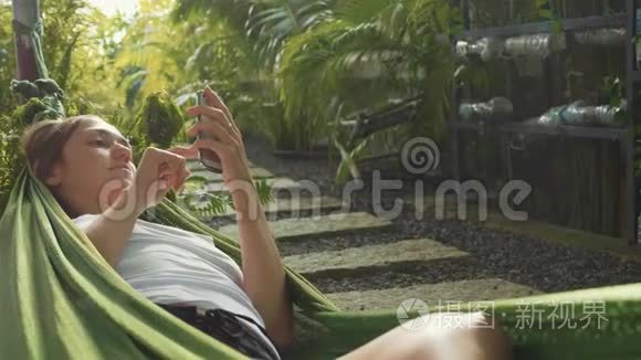 年轻女子躺在绿色花园的吊床上使用智能手机。