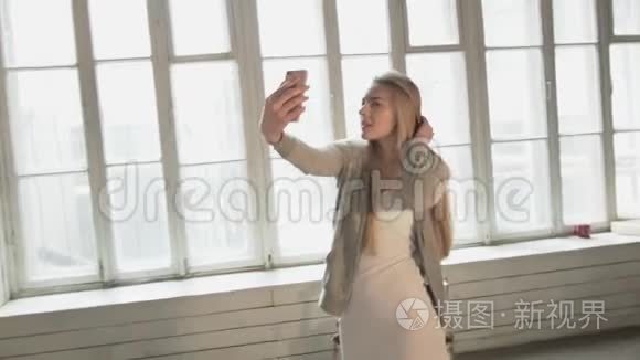快乐美丽的年轻女人在靠窗的地方自拍。 带着智能手机笑的金发美女给自己拍照。