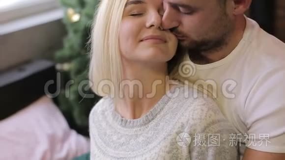 年轻夫妇坐在圣诞树背景上的床上。 在家过喜庆的夜晚。 男人亲吻女人