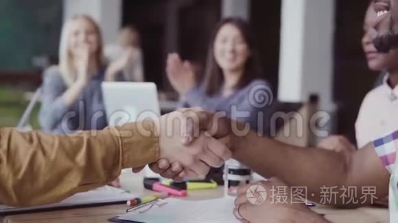 两个伙伴，非洲和高加索商人握手。 一群人在现代办公室的背景上鼓掌。