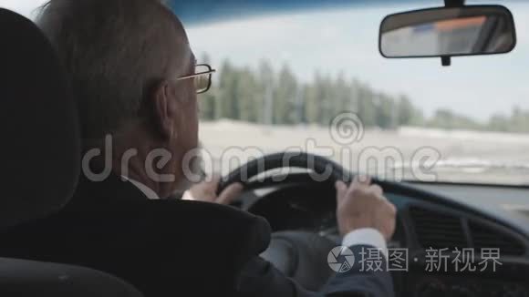 一个头发灰白，夹克里有胡子的男人坐在汽车的车轮后面。