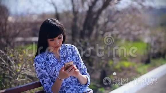 美丽的亚洲女孩坐在公园里的长凳上，在智能手机上读着一条信息，非常惊讶