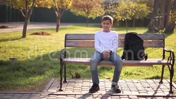 穿白色毛衣的少年坐在长凳上。 带背包的帅哥学校男孩
