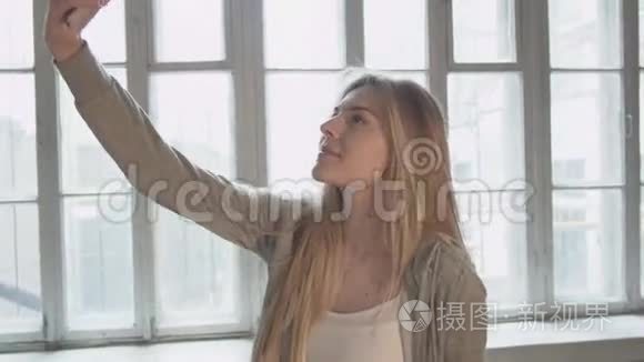赛尔夫。 年轻漂亮的金发女郎拿着一部手机对着一扇大窗户。 送一个空气吻。 在一个