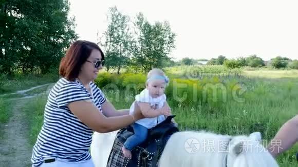 一个带着孩子的女人在田野里走来走去，一个婴儿坐在小马上，妈妈抱着孩子。 快乐的家庭假期