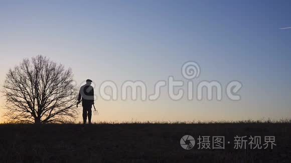 一个拿着铲子在田里散步的农夫。 他们携带种植树木的设备。 在日落的时候。 侧视图
