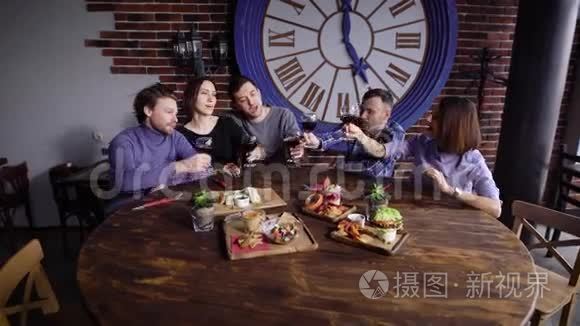 一群可爱的年轻人在一家现代化的的桌子上吃零食。 男人和女孩用酒杯烘焙