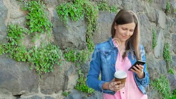穿着棉袄用智能手机喝咖啡的漂亮女孩。 背景中野石的复古墙