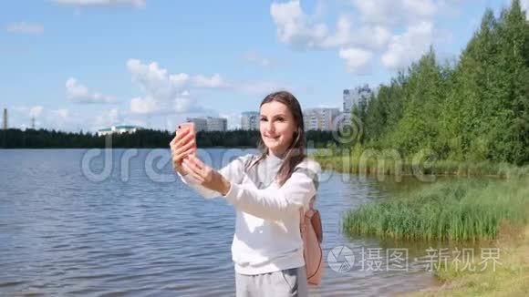 带背包的漂亮女孩在湖边自拍，并在Skype上用手机交谈。