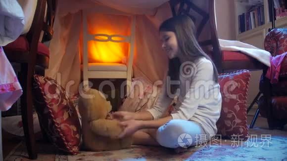 多莉拍了一张可爱的笑脸女孩，穿着睡衣，在用毯子做的房子里和泰迪熊玩耍