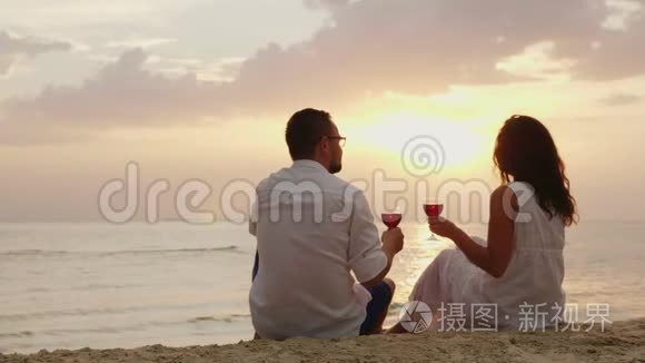 一对年轻夫妇在日落时正在海边喝酒。 他们坐在沙滩上，戴着眼镜。 周年或蜜月