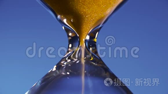 透明烧瓶中的玻璃金色沙子倒在蓝色的背景上，点缀着时间的短暂