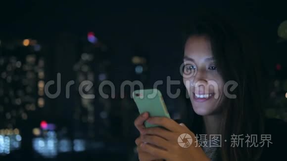 年轻的微笑女人在城市的夜晚在手机上发短信。 4K. 使用智能手机的混血女孩