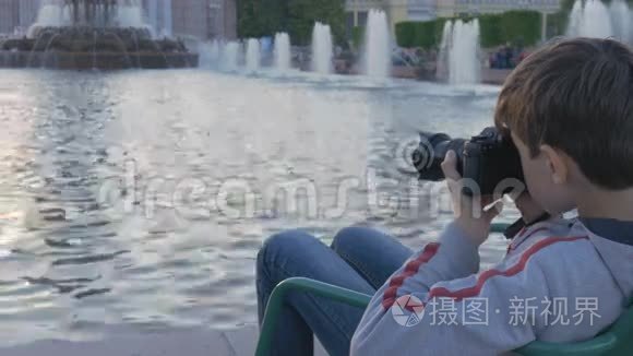 这个男孩是个十几岁的孩子，坐在喷泉旁边的一个美丽的地方，拍摄水溅的照片。 美丽的风景