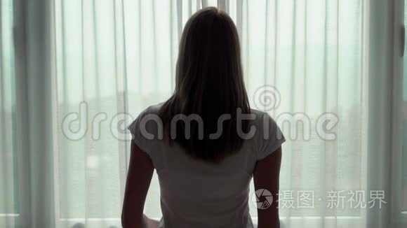 年轻女人揭开窗帘的剪影，望着窗外。 在外面欣赏海景。 慢动作100fps..